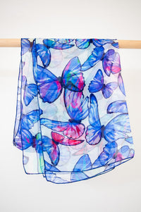 A large silk scarf with a violet butterfly pattern, displayed on a wooden rod with a white background, front view