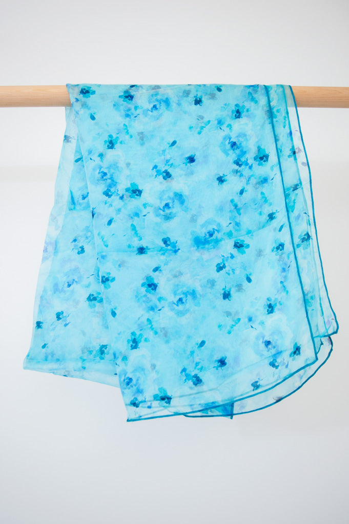 A blue silk scarf with a watercolour floral pattern displayed on a wooden rod against a white background, front view