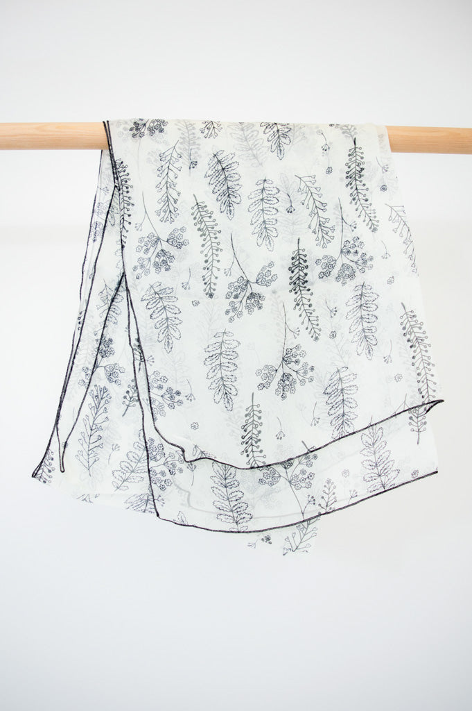 A large silk scarf with a black and white leaf pattern displayed on a wooden rod against a white wall, front view