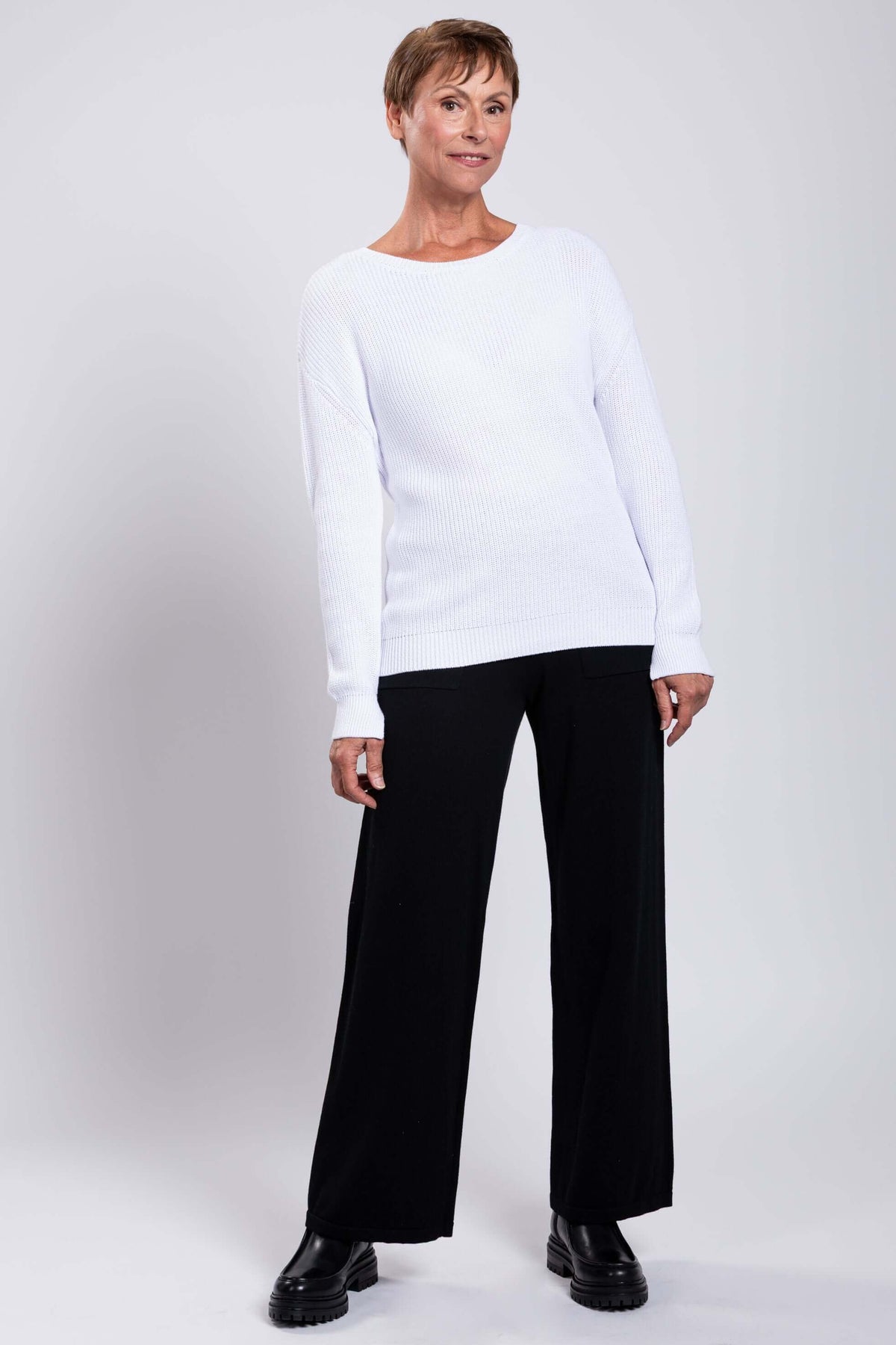 Model in a white cotton sweater and black pants standing with one hip tilted