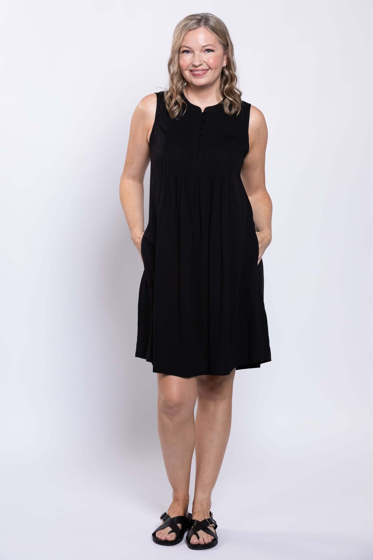 Woman smiling in sleeveless black A-line dress with pintucked front, hands in pockets