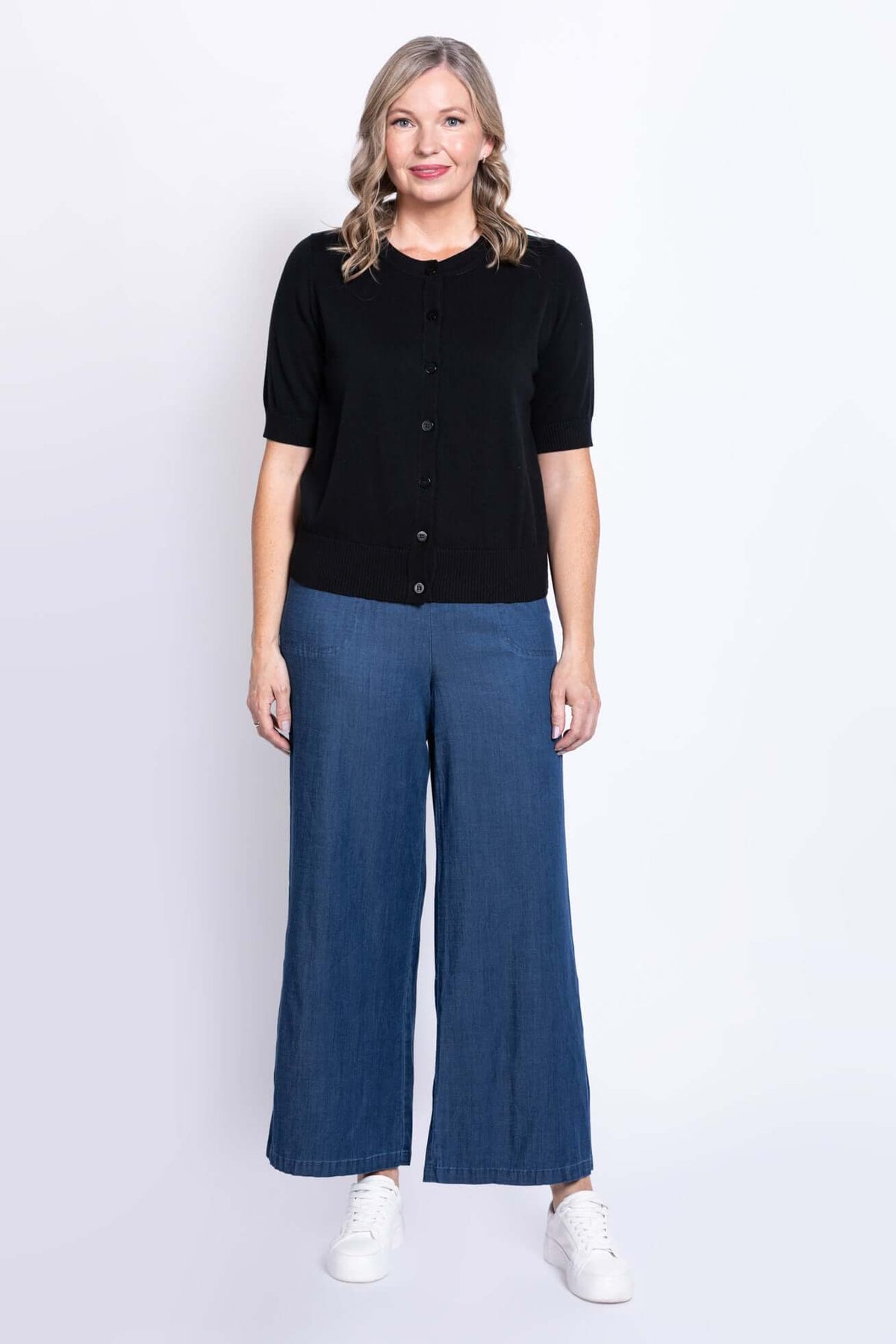 Woman facing forward in a black button-up short-sleeve cardigan, blue wide-leg pants, and white shoes