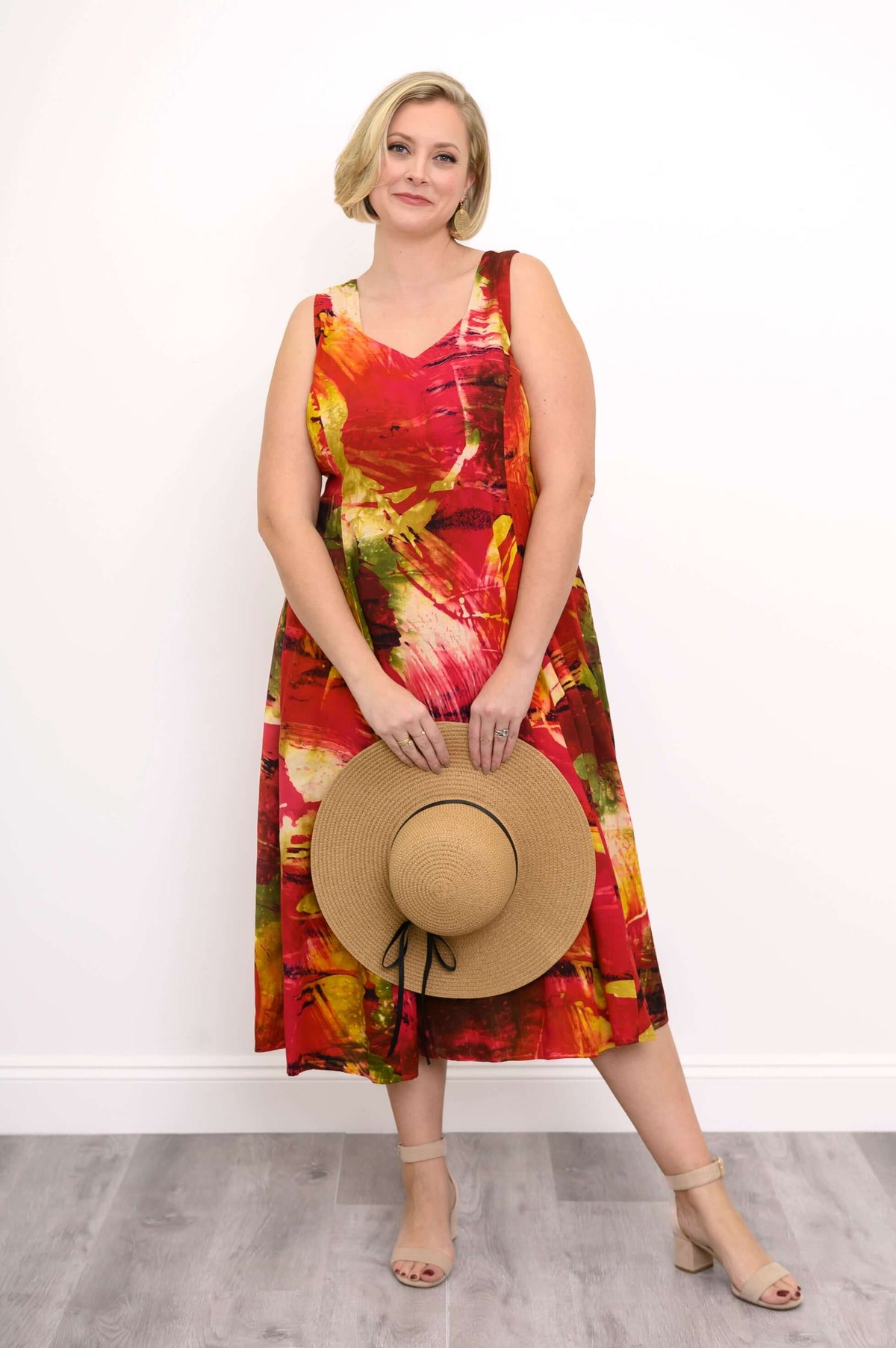 Woman in a colorful sleeveless midi dress holding a straw hat in front, standing on a wood floor
