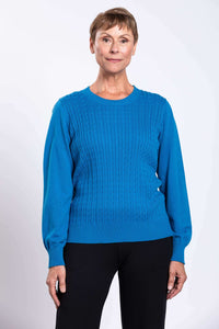 Woman wearing a bright blue cable knit crew neck sweater with black pants