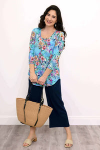 Full body view: woman in blue floral bamboo top, navy pants, holding a straw tote bag