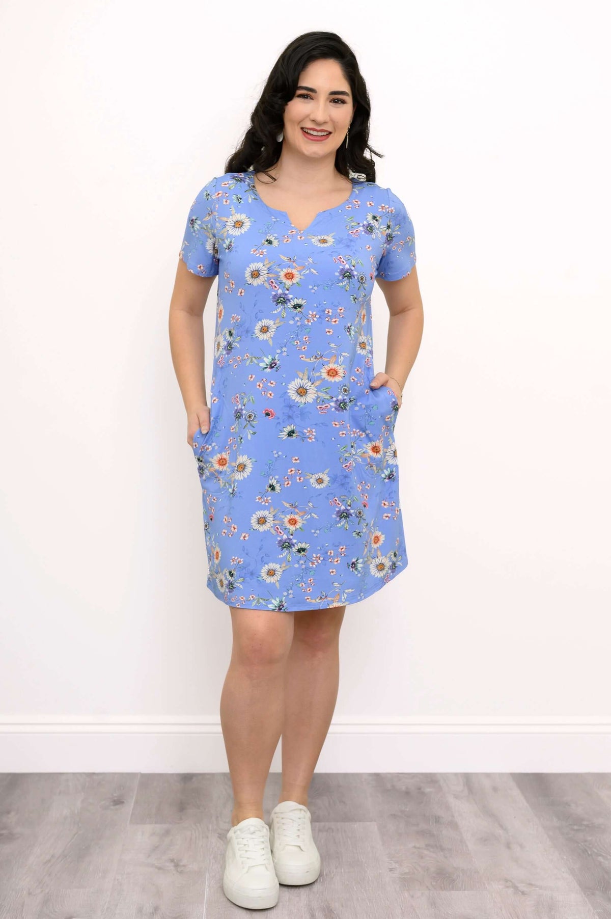 Woman in blue floral shift dress standing with hands in pockets and white sneakers