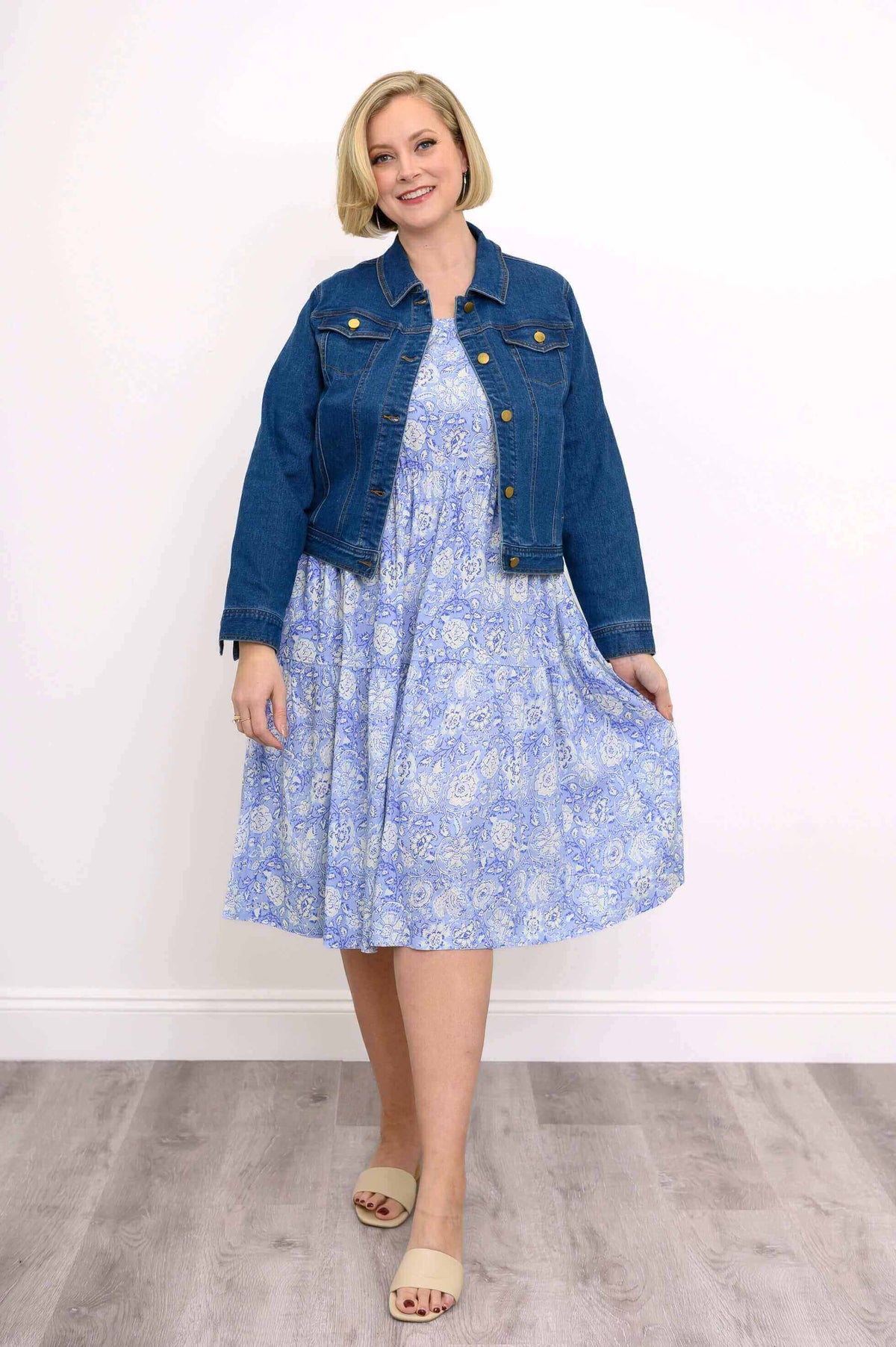 Woman wearing a blue floral tiered dress with a cropped denim jacket and beige sandals