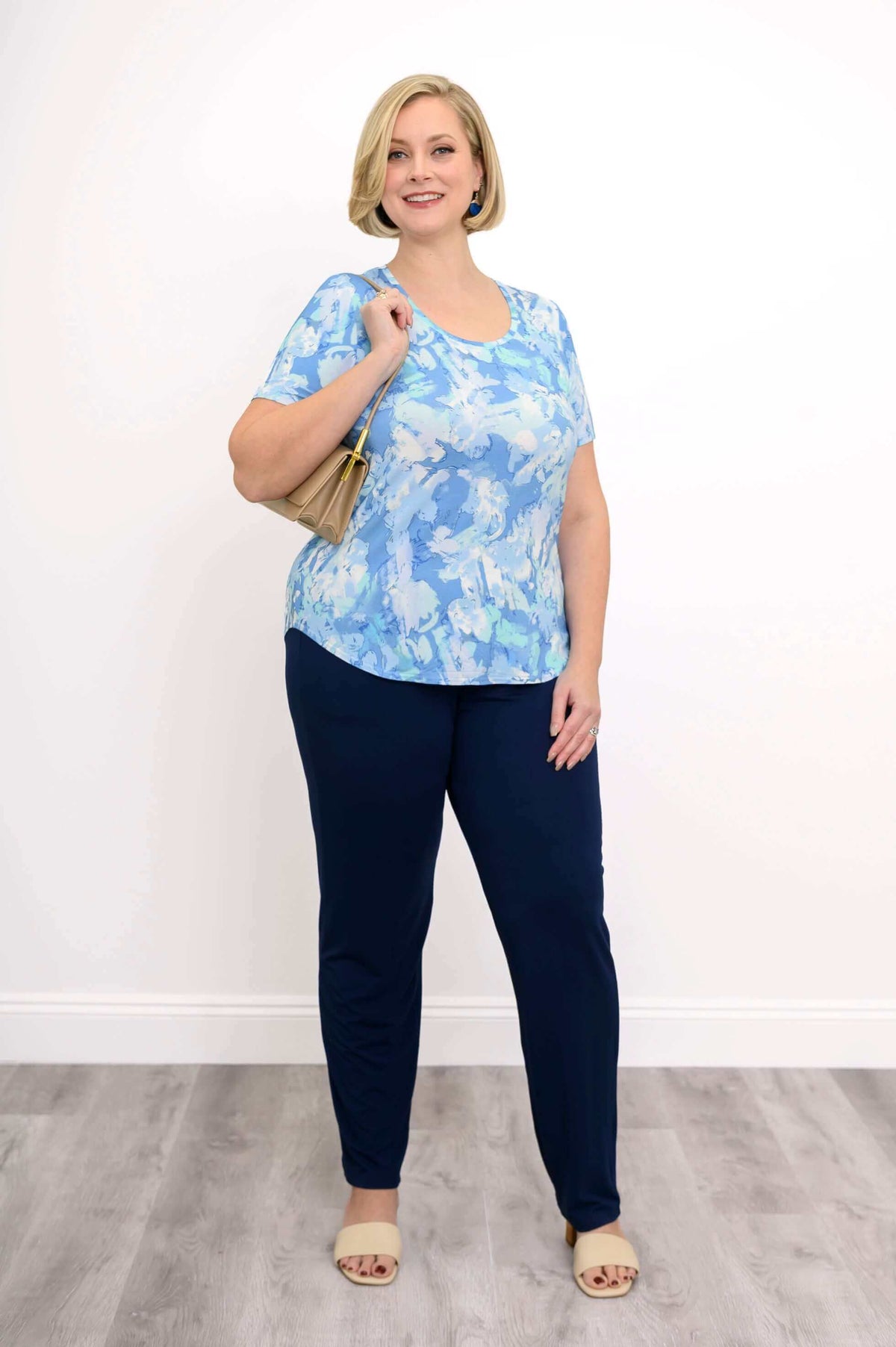 Smiling woman in a blue watercolor tee and navy pants holding a tan handbag