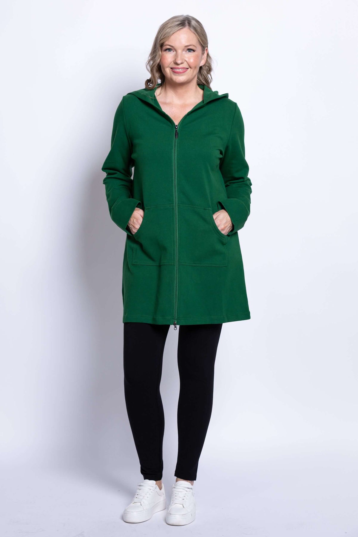 Woman in a green hooded jacket, hands in pockets, black leggings, and white shoes