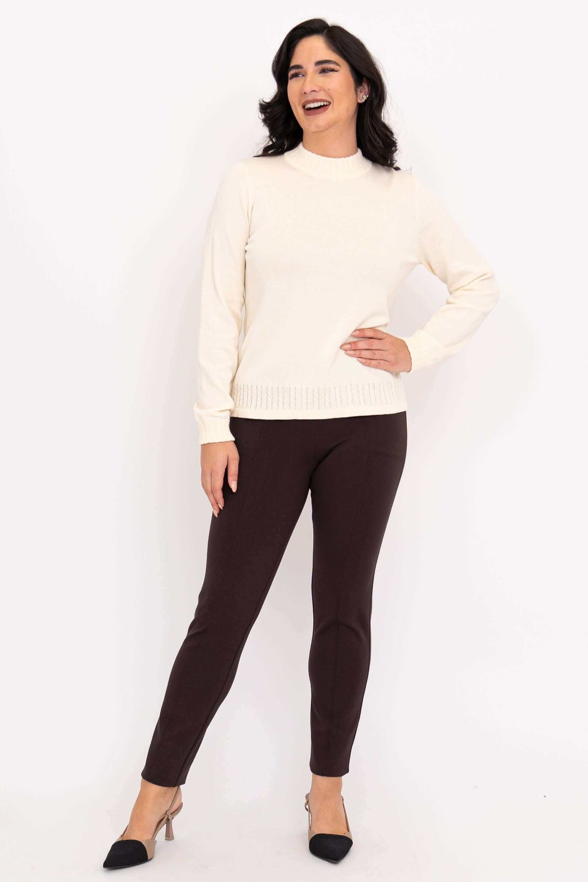 A cheerful model wearing an off white cable trim knit sweater paired with dark slim leg pants.