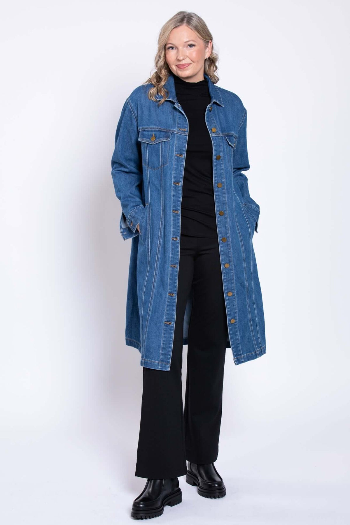 Woman in long denim jacket, hands in pockets, black pants and boots, white background