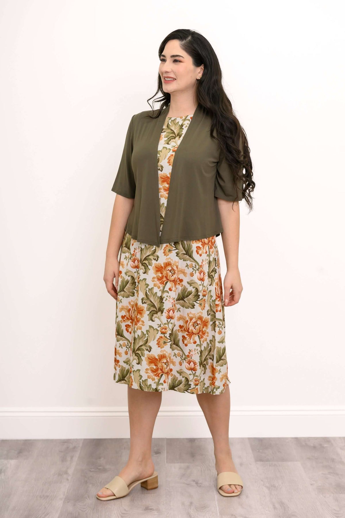 Woman in olive shrug over floral sleeveless dress posing sideways