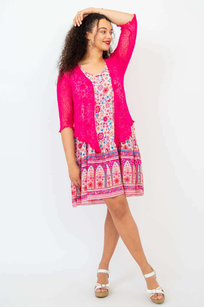 Woman in a pink floral dress paired with a bright pink lace cardigan, posing with one arm raised.