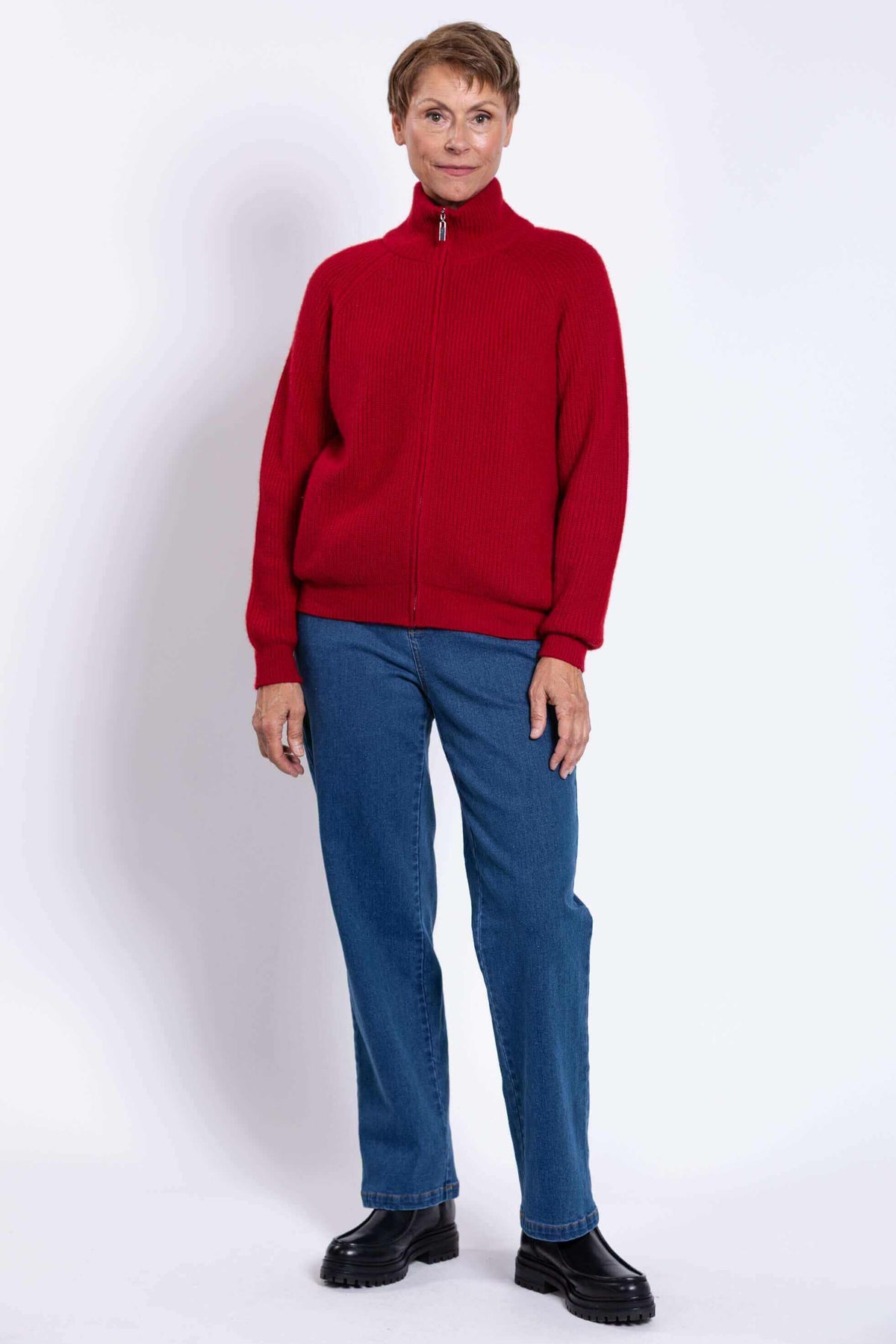 Woman in a red cardigan sweater, blue jeans, and black boots, posing against white background