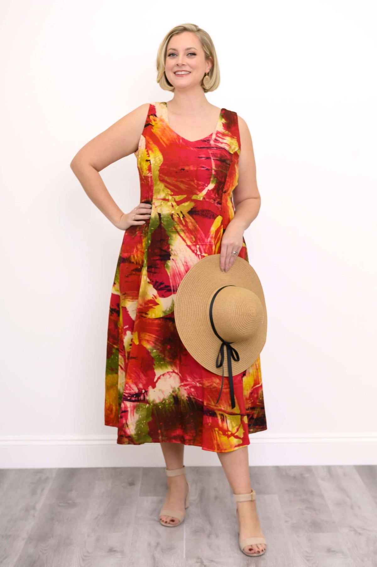Woman wearing a sleeveless midi dress with a vibrant red, yellow, and green abstract print, holding a straw hat