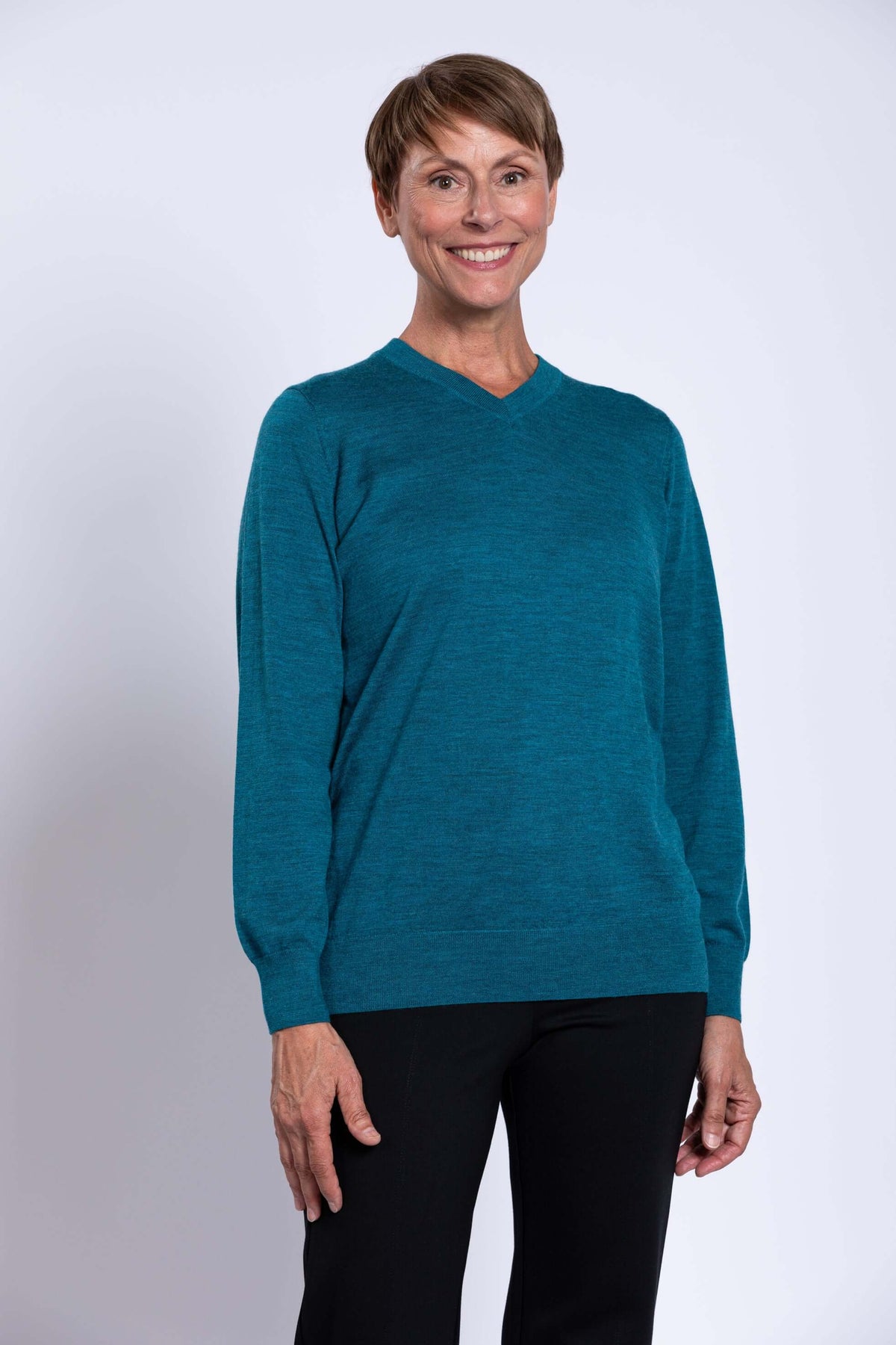 Woman wearing a teal V-neck Merino wool sweater with black pants, smiling in front of a white background