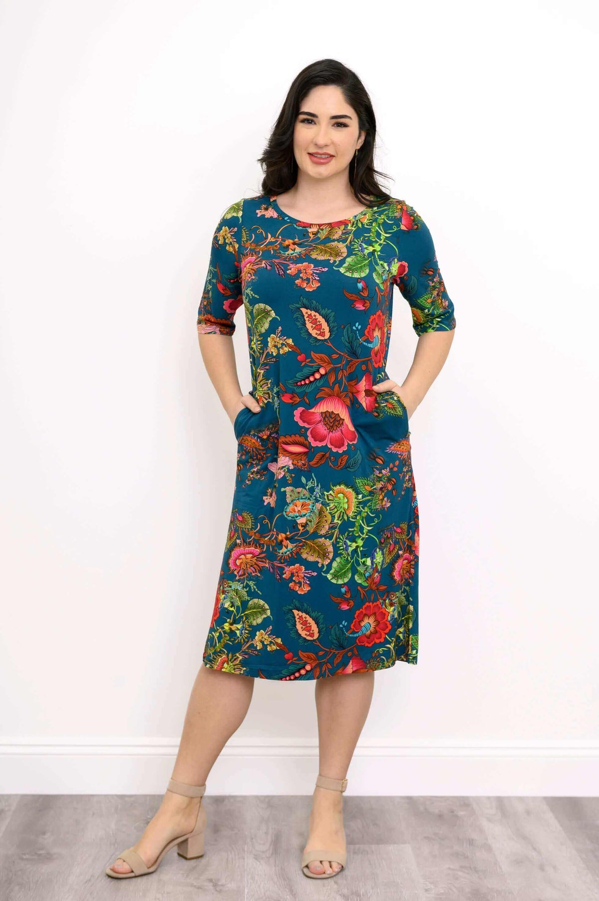 Woman posing hands in pockets, wearing teal floral midi dress with bright flowers