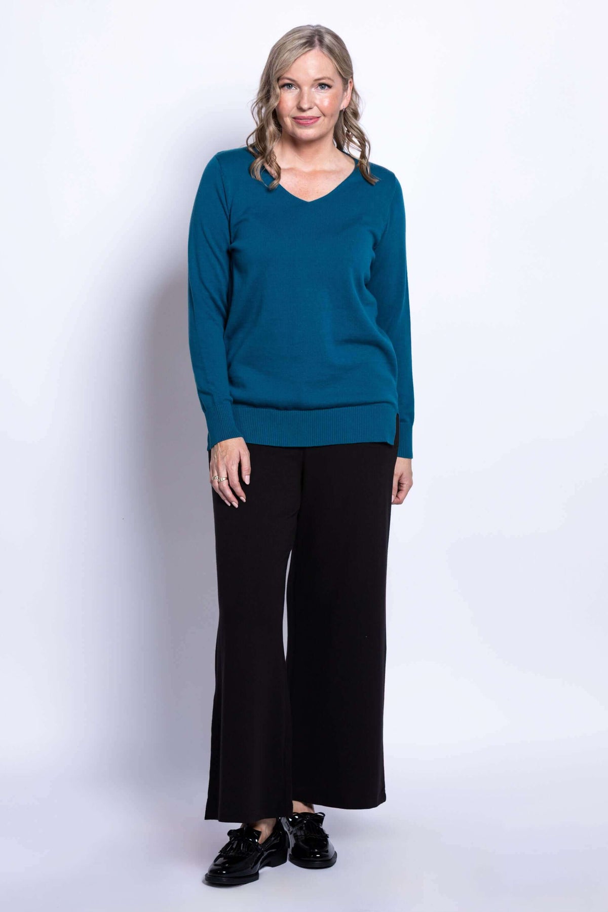 Woman standing in teal v-neck sweater and black pants against white background
