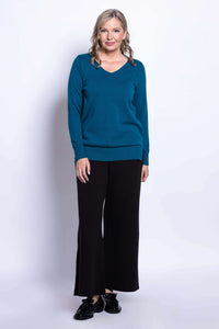 Woman standing in teal v-neck sweater and black pants against white background