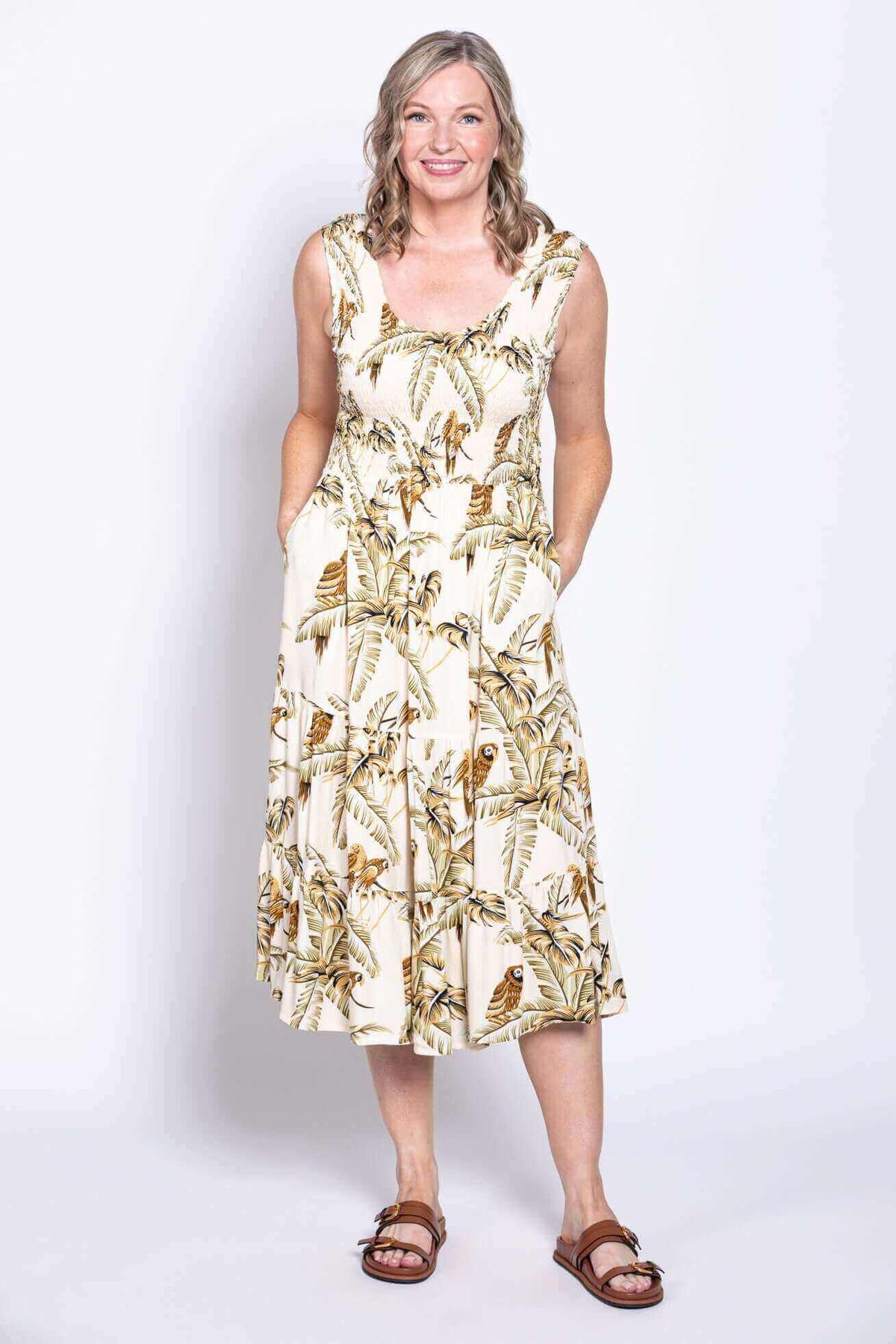 Woman in sleeveless tropical print midi dress with hands in pockets, smiling front facing