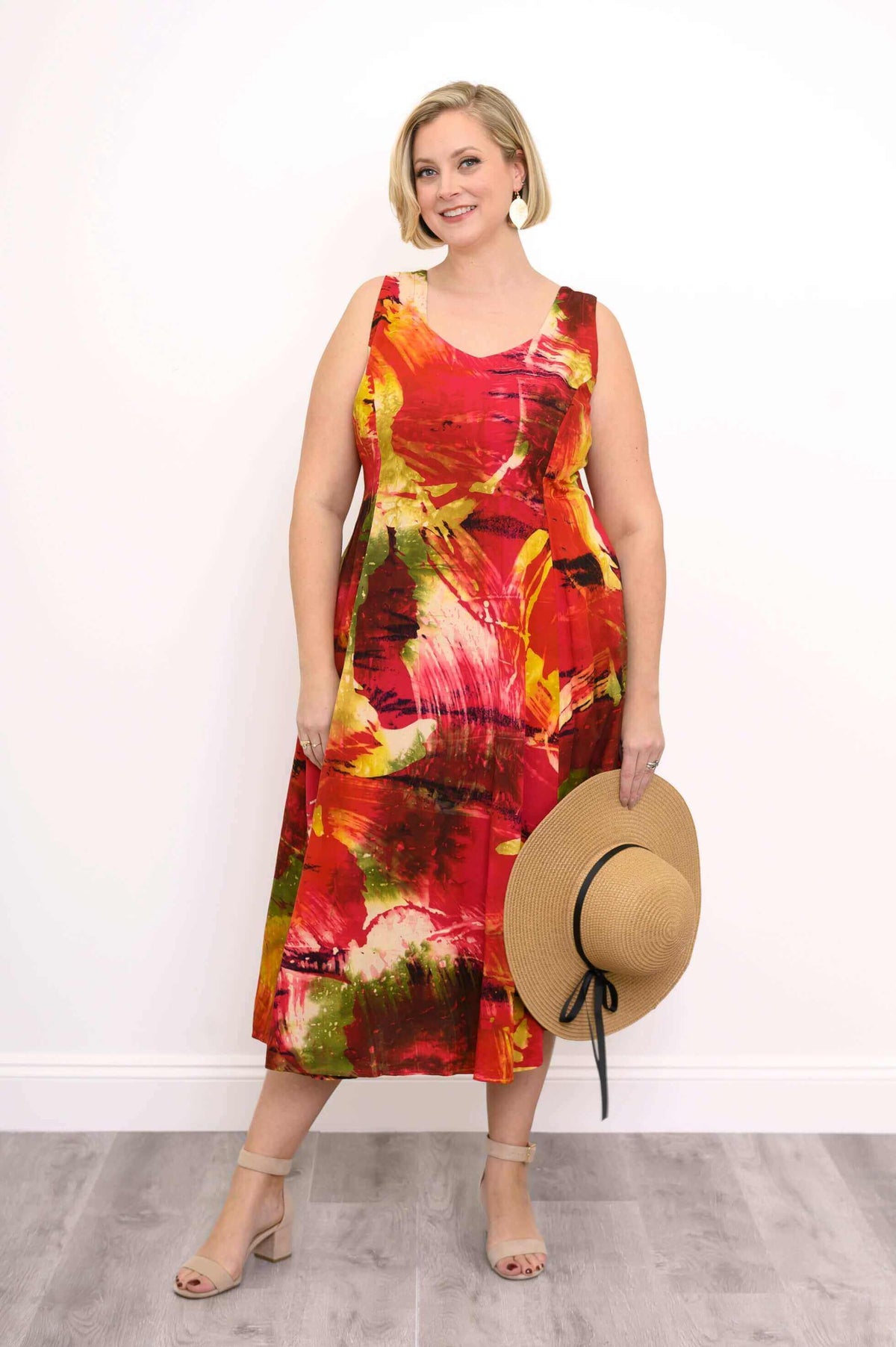 Woman in a sunset-inspired sleeveless midi dress holding a straw sunhat, smiling at the camera