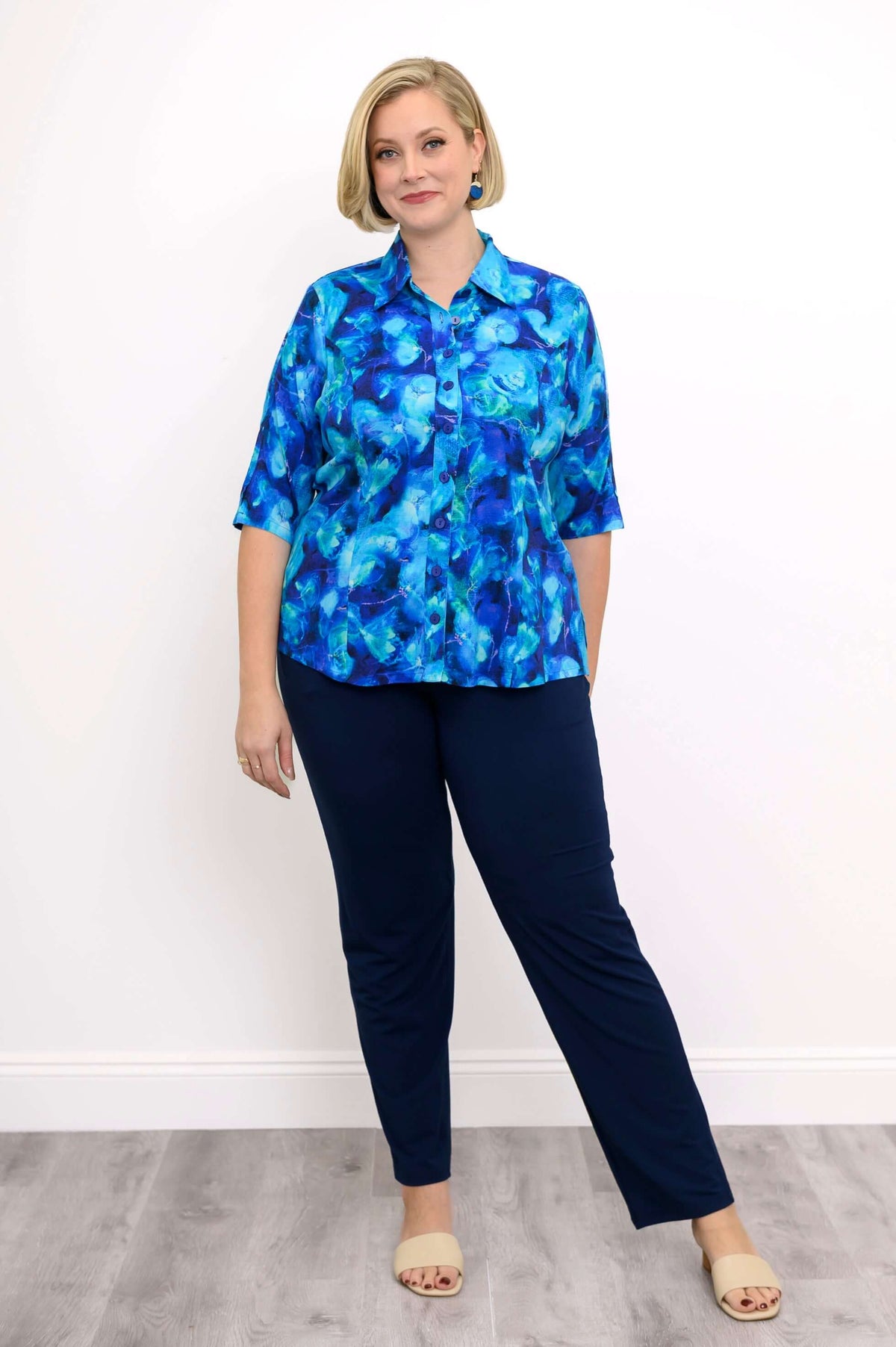 Woman in watercolor blue blouse with navy pants, standing against white wall