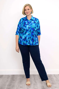 Woman in watercolor blue blouse with navy pants, standing against white wall
