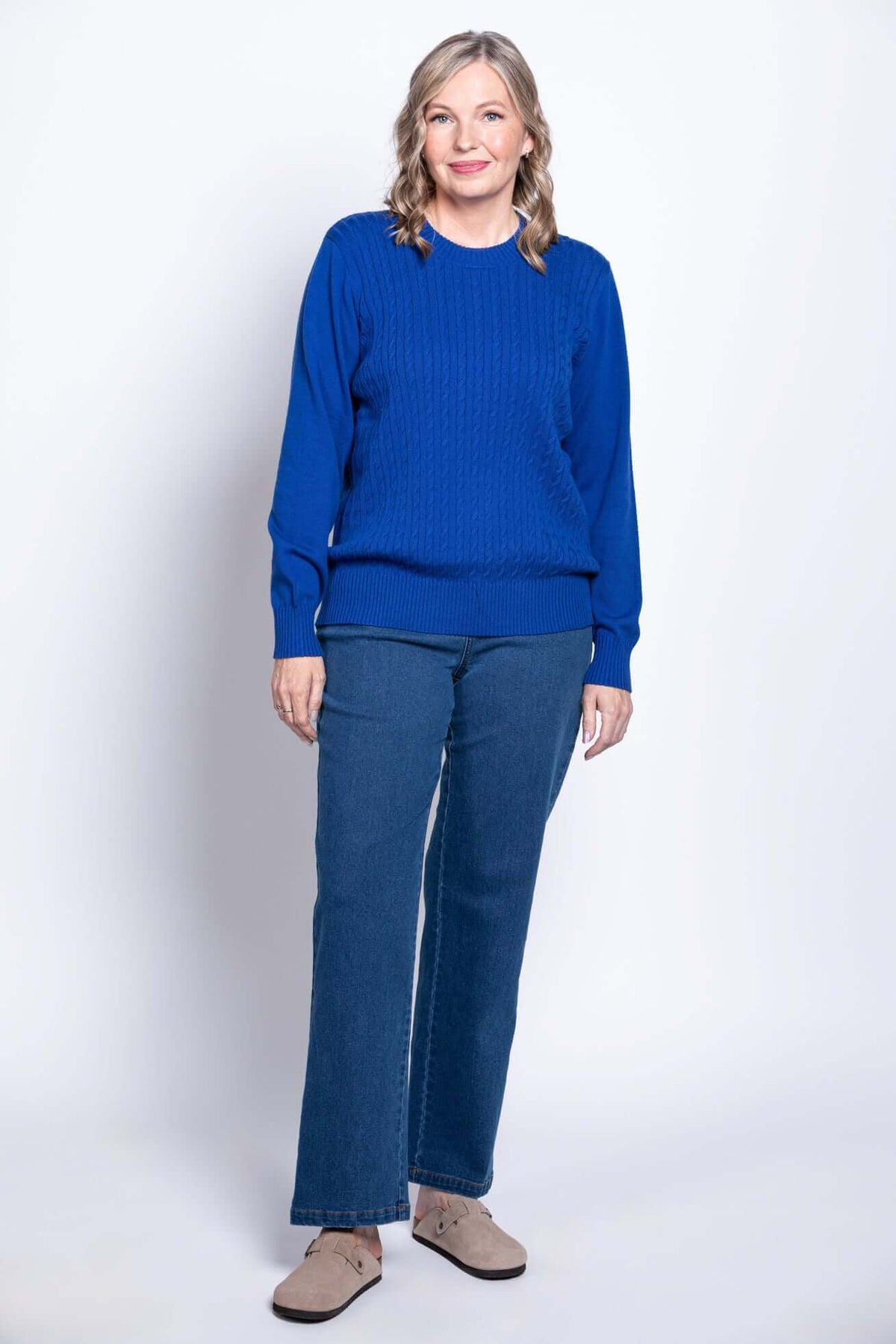 Full length shot of a woman in a royal blue cable knit sweater, dark blue jeans, and beige clogs.