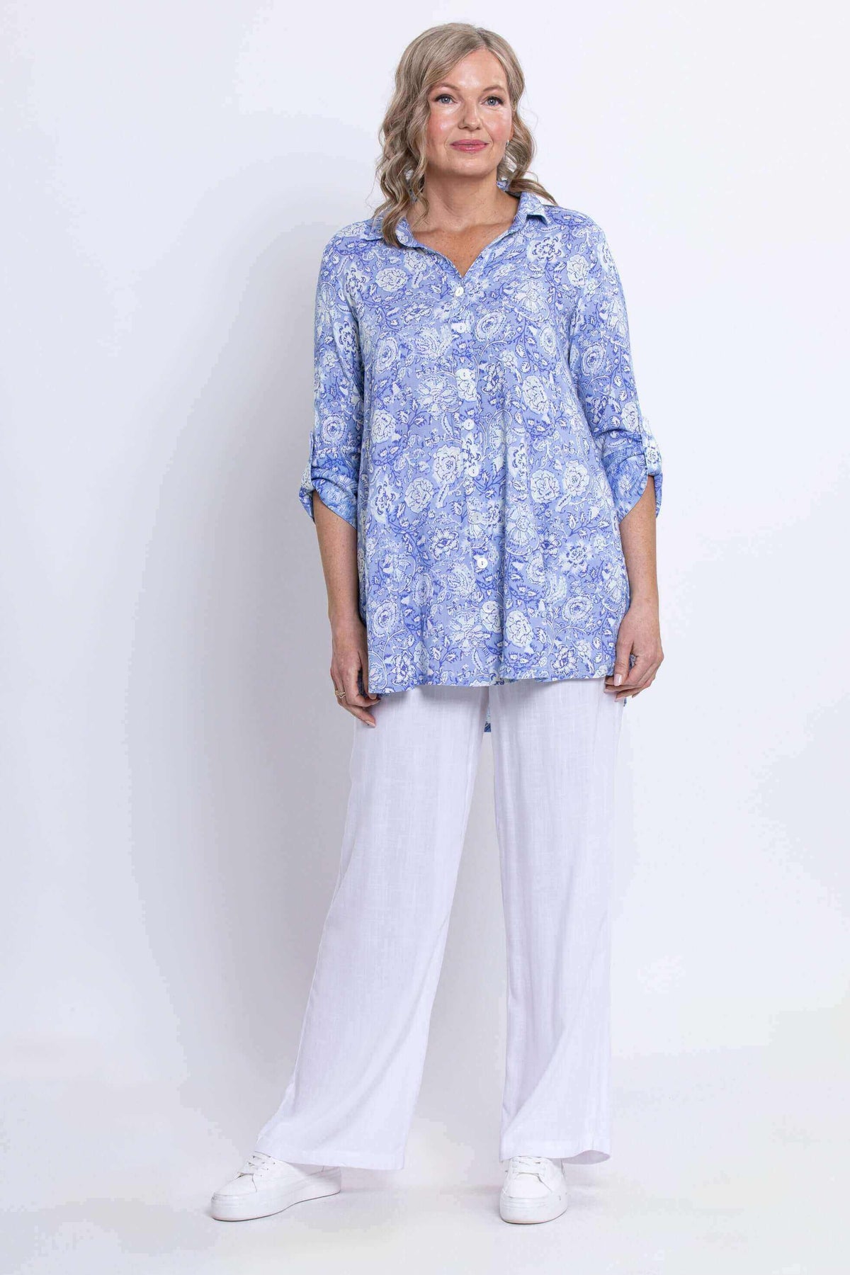 Woman posing in blue paisley tunic and white pants, hands in pockets