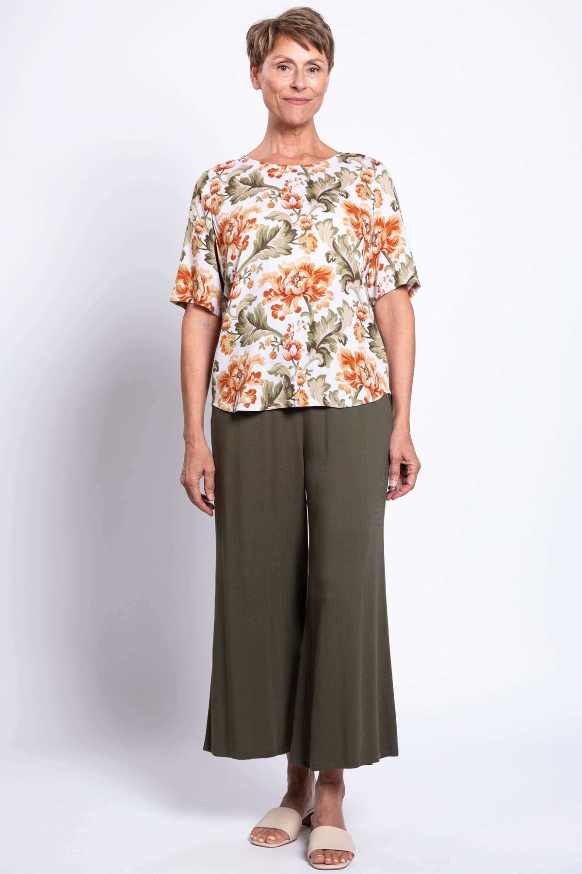 Woman standing in floral blouse, olive pants, sandals, facing forward