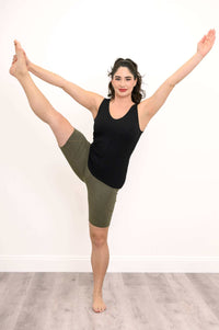 Woman in olive green bike shorts and black tank top, doing a standing split yoga pose