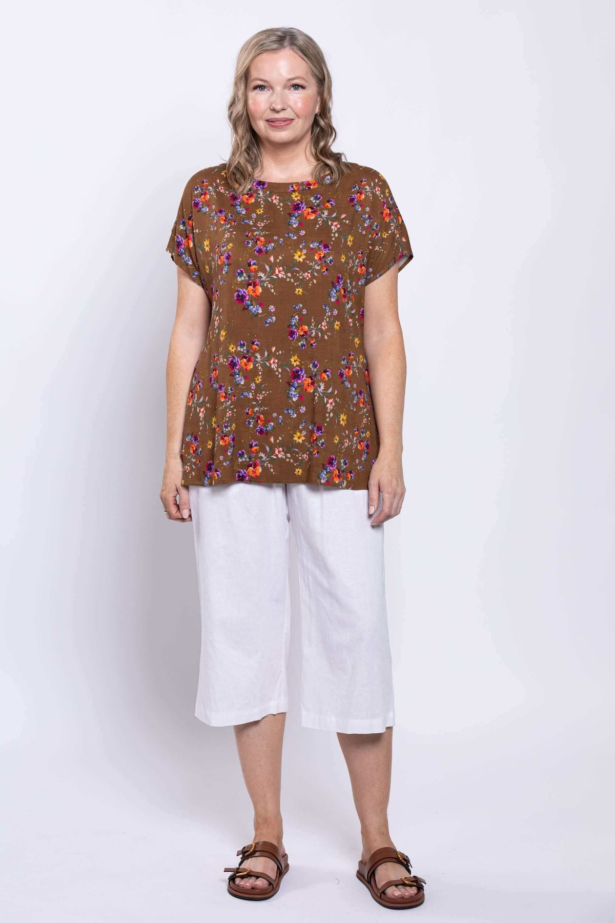 Woman standing in brown floral tee with colorful floral print, white pants and sandals