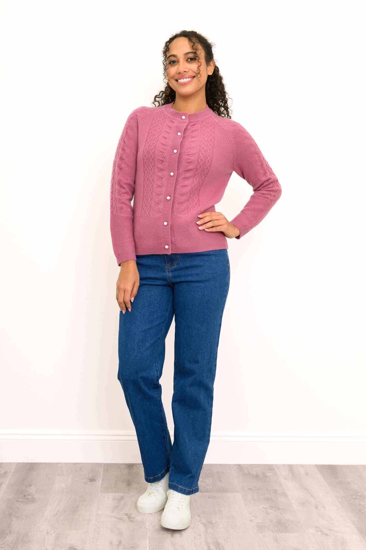 Woman posing in a pink cable knit cashmere cardigan, blue jeans, and white sneakers.