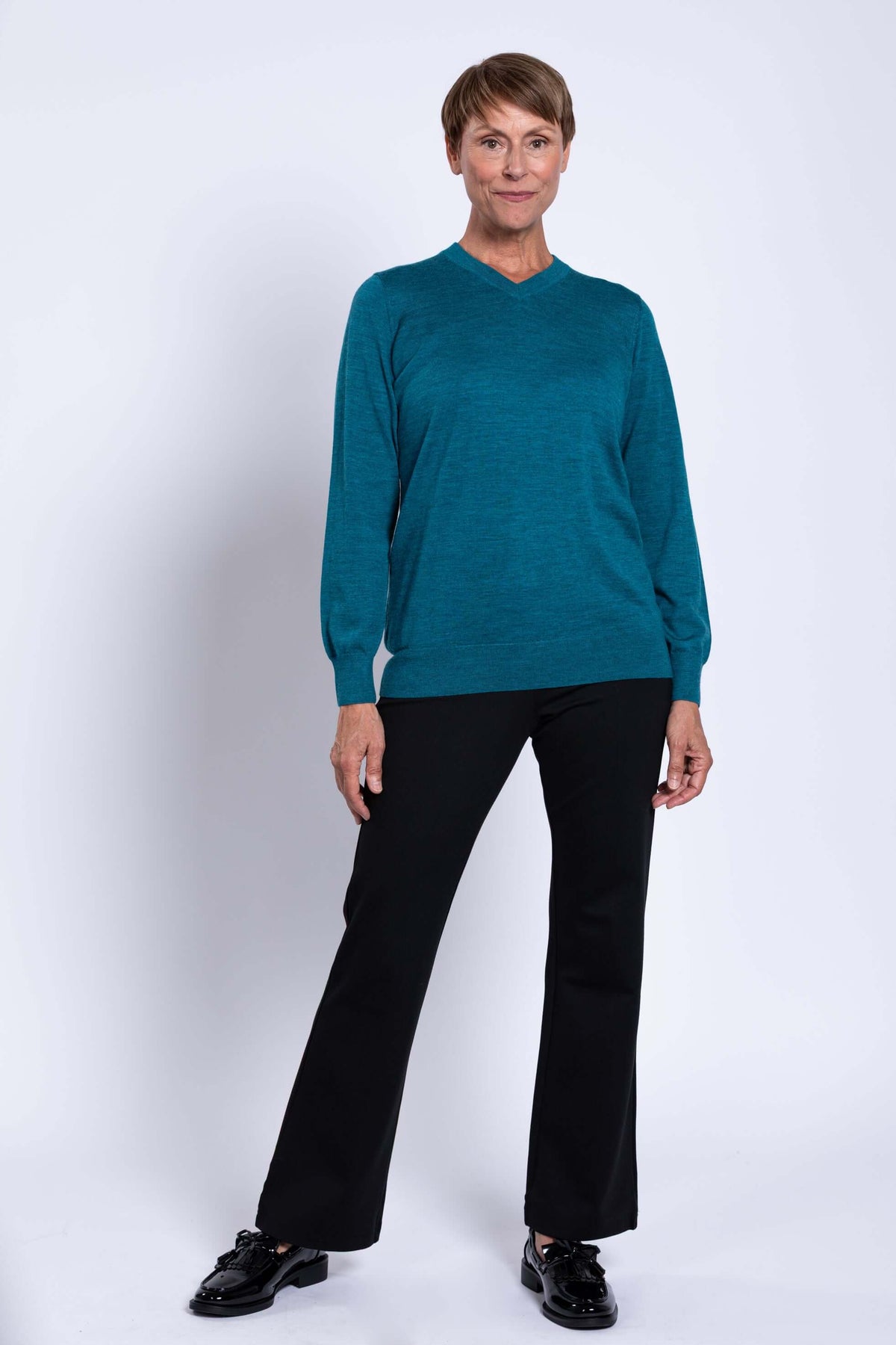 Woman posing in a teal V-neck Merino wool sweater and black pants, with one leg slightly forward