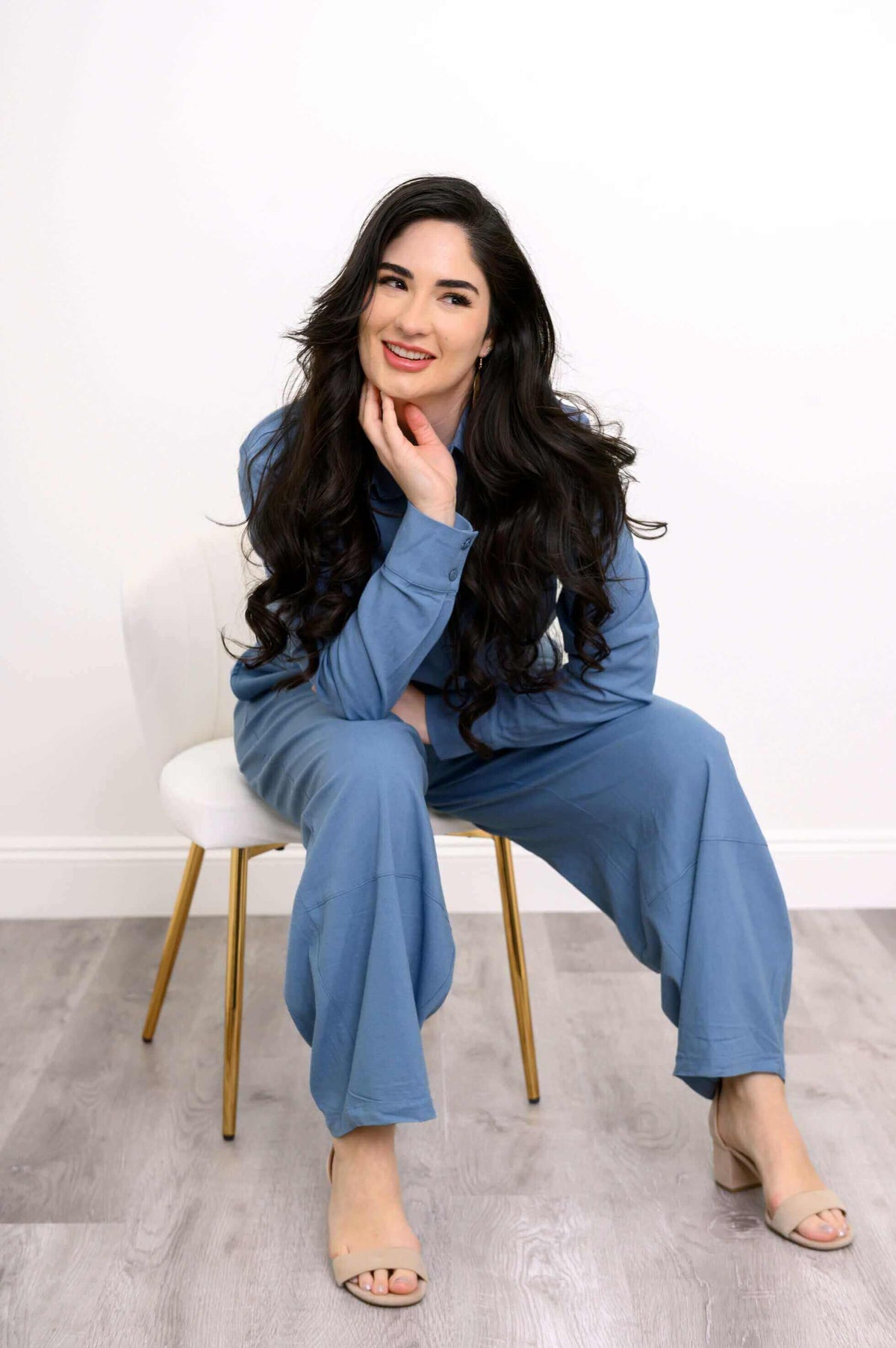 Smiling woman in blue shirt and pants set, seated on a white chair, posing casually