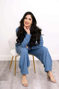 Smiling woman in blue shirt and pants set, seated on a white chair, posing casually