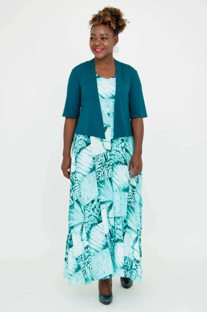 Woman smiling and modeling teal cropped Doda Jacket over green patterned dress