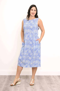 Woman smiling and standing with hands in pockets in a blue floral sleeveless dress