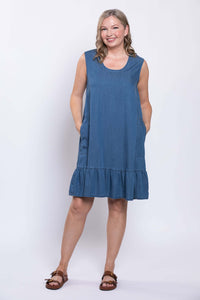 Woman in blue sleeveless dress with ruffle hem standing hands in pockets and smiling