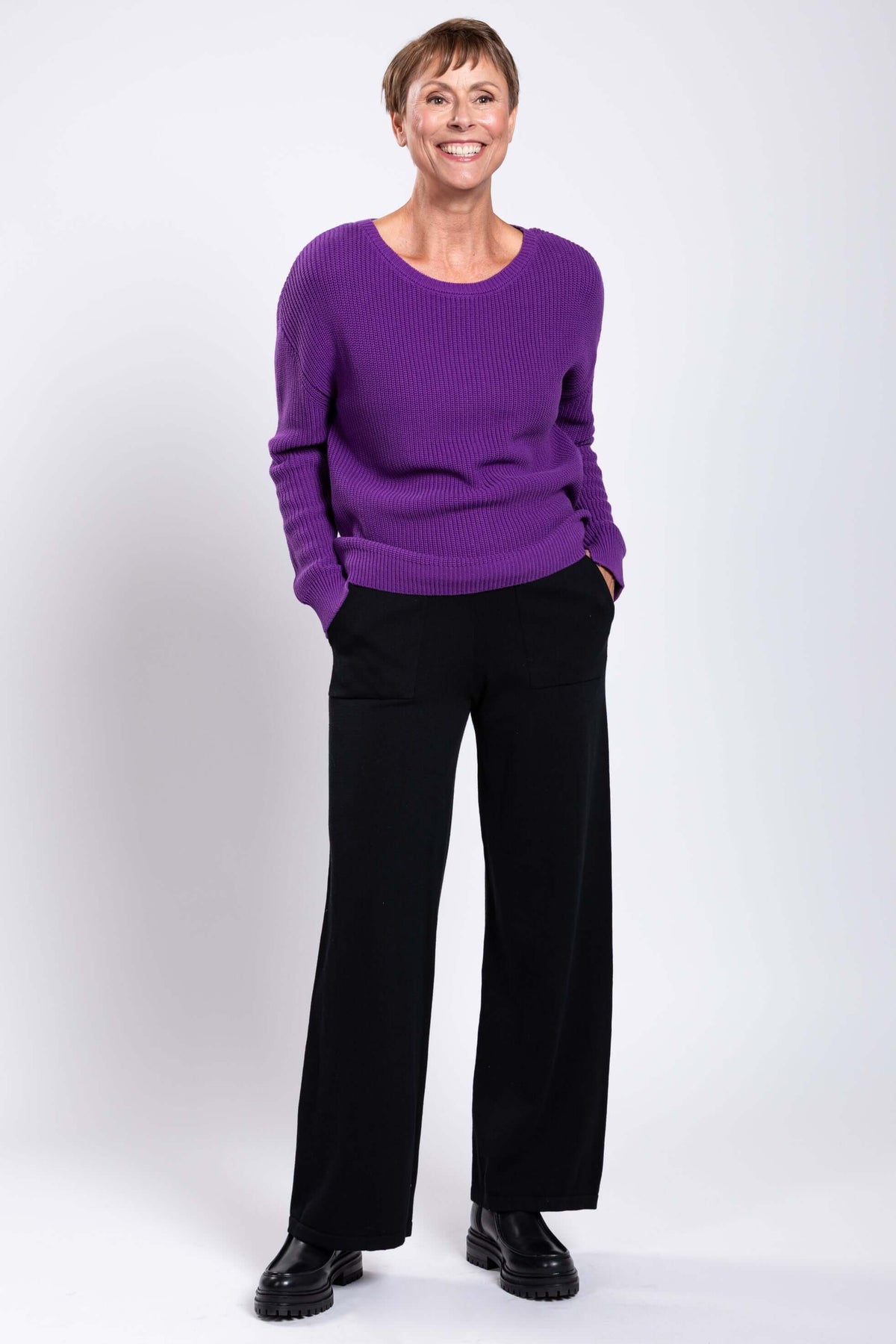 Woman smiling while wearing a purple shaker stitch sweater and black pants