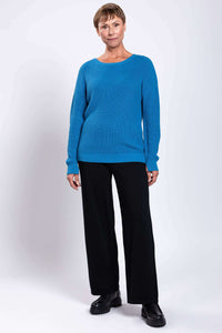 Woman standing in a blue knit sweater and black pants, facing forward