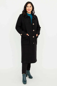 Woman standing in a knee-length black coat with hands in patch pockets