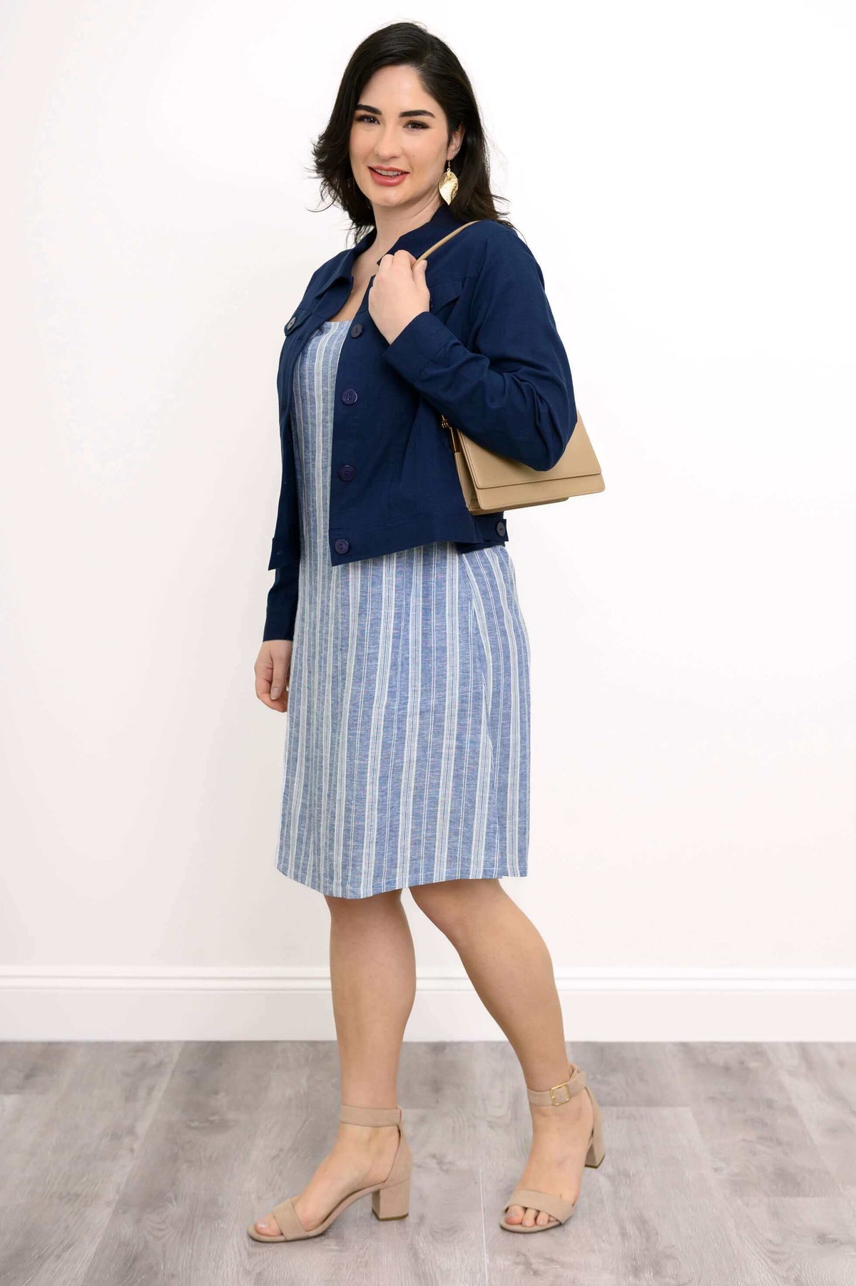 Woman in blue jacket and striped shift dress holding a beige handbag.