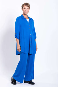 Side angle of a woman in a blue blouse-jacket and matching pants, hands in pockets