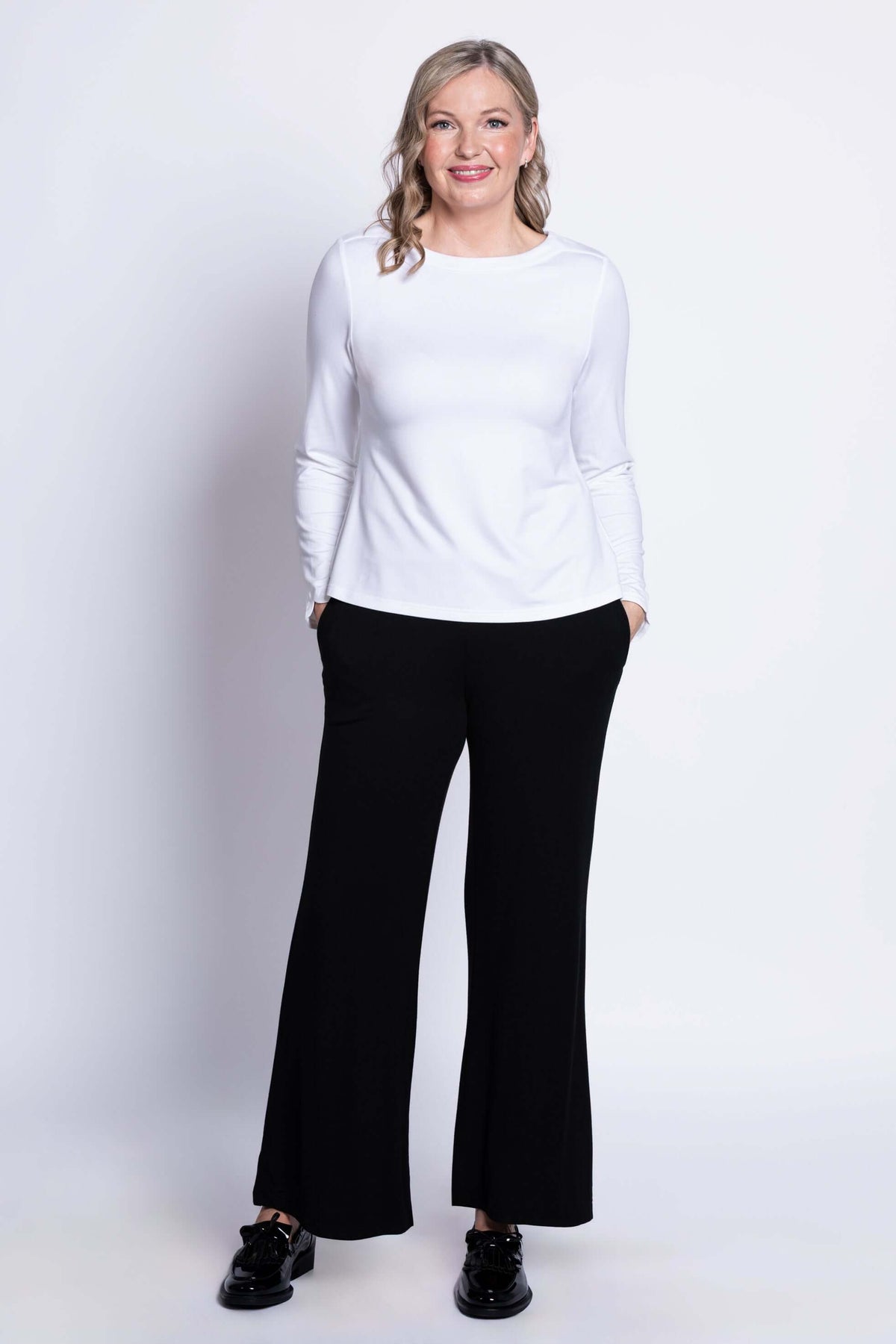 Woman with hands in pockets wearing white top and black pants, front pose